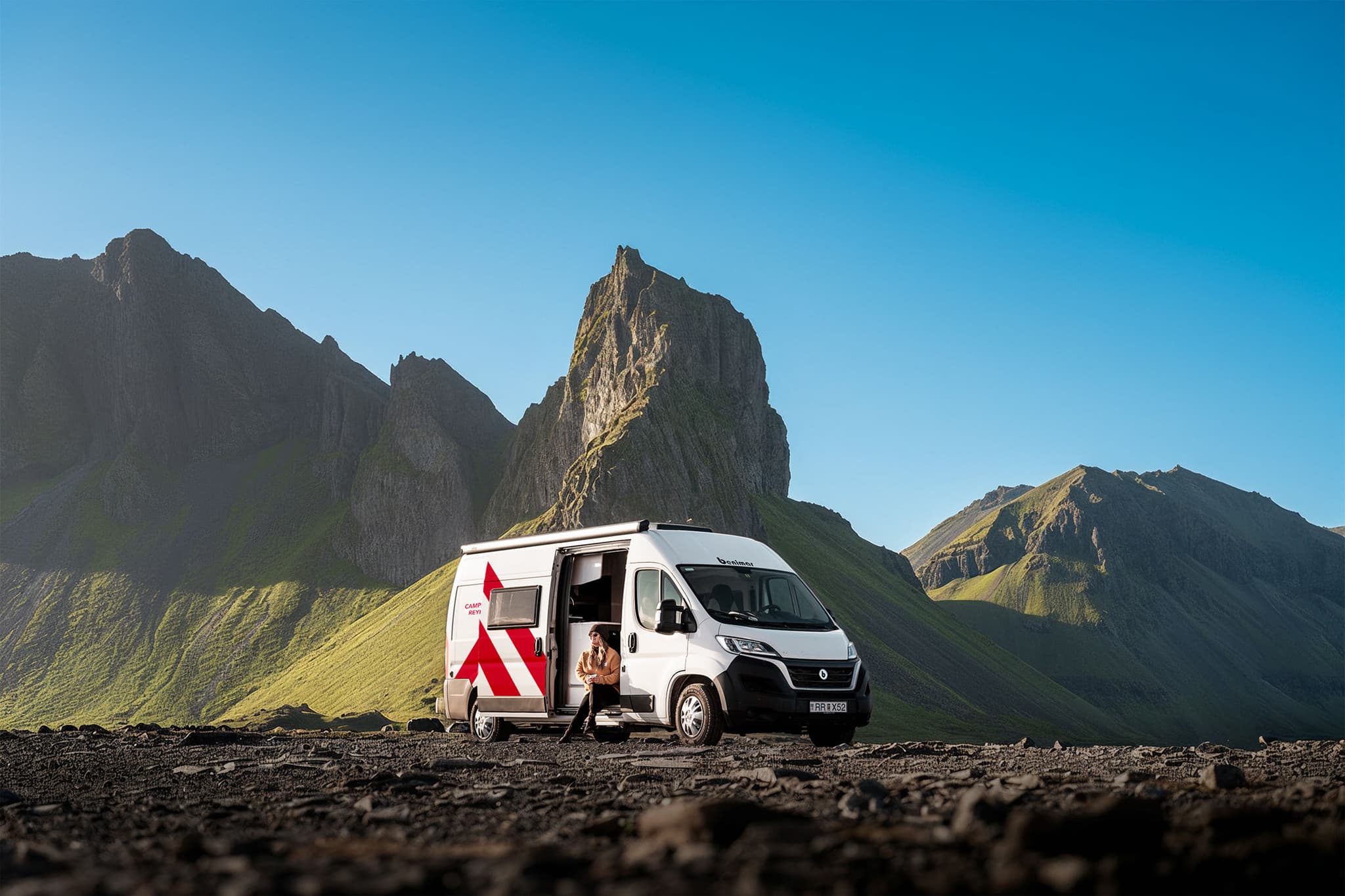 Campervan Rentals in Iceland - Free Extras, Unlimited Mileage & Flexible Cancellation | Campervan Reykjavik White Campervan Reykjavik rental on black gravel, side door open with traveler seated; green volcanic peaks and clear summer sky in Iceland.