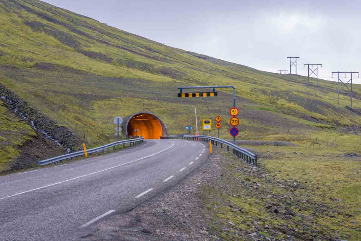 Road signs and clearance bar at the approach to an illuminated tunnel on an Icelandic mountain pass.