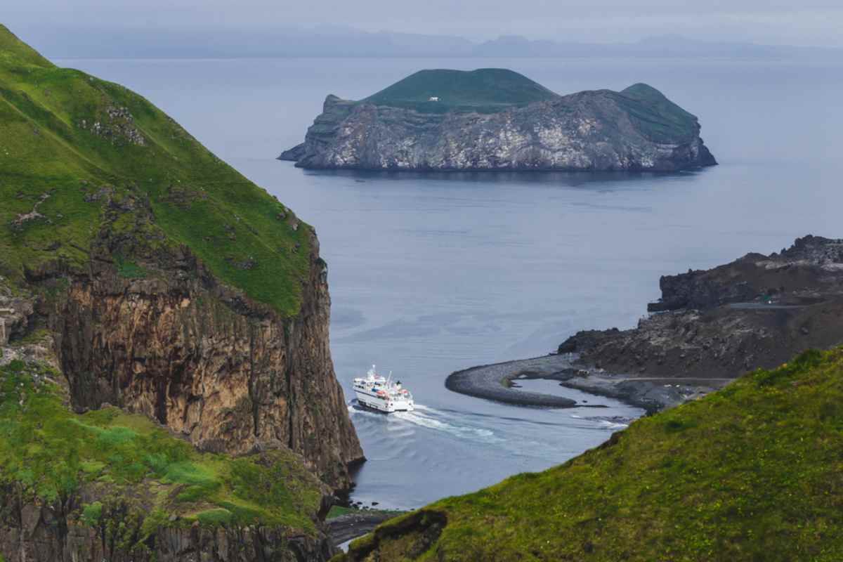 Ferry navigating a narrow channel between steep green cliffs toward an offshore island in calm seas.