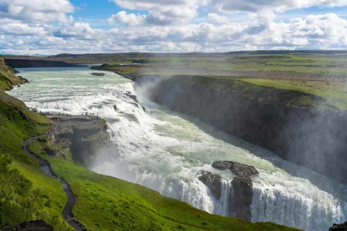 Large waterfall in Iceland tumbling between green hills under a partly cloudy sky.